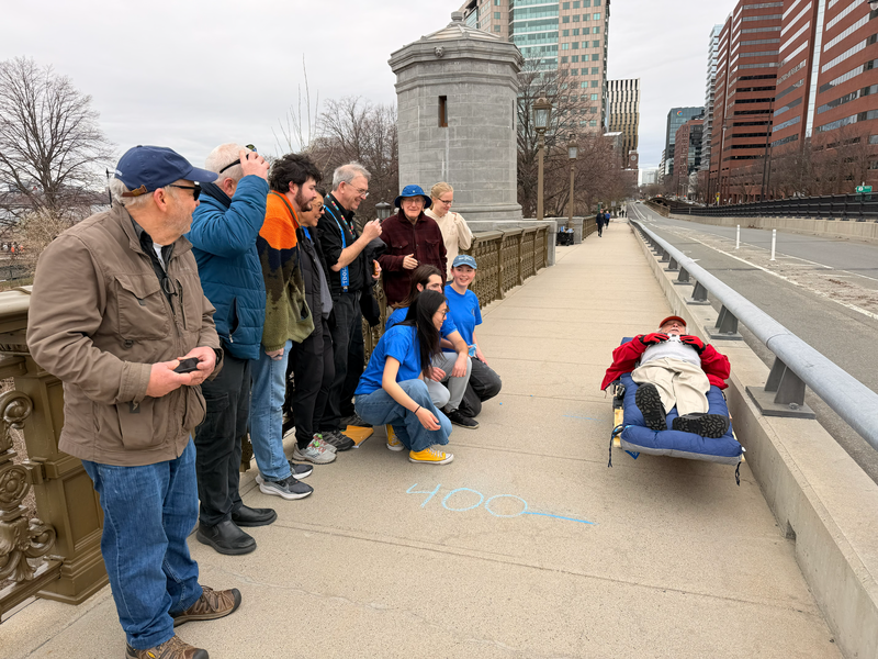 Ten people stand and kneel on a bridge sidewalk next to Martin Kelin, who is laying down on a wooden cart.
