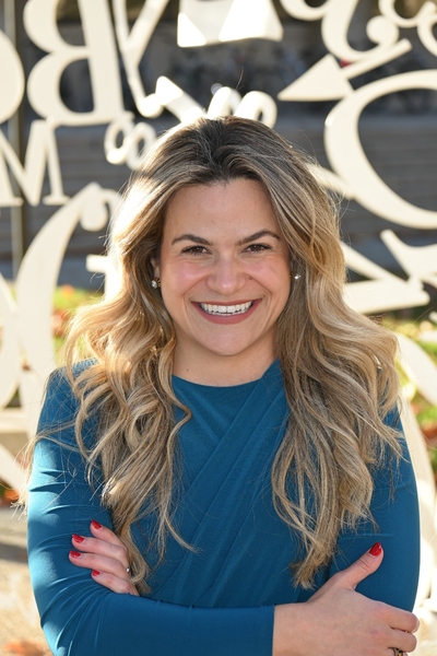 A woman in a light blue shirt smiles with her arms crossed in front of Alchemist, a large outdoor sculpture on the MIT campus.