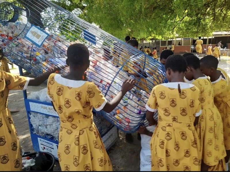 High school students in yellow uniform dresses handle a large wire basket filled with plastic waste