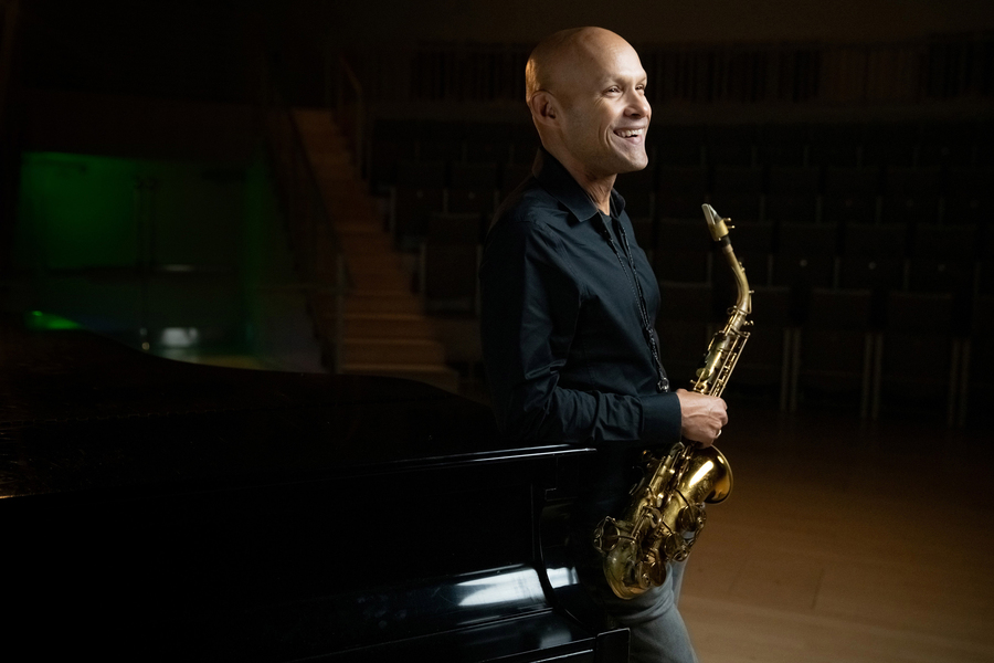 Miguel Zenón leans against piano and holds the saxophone in a dark music room.
