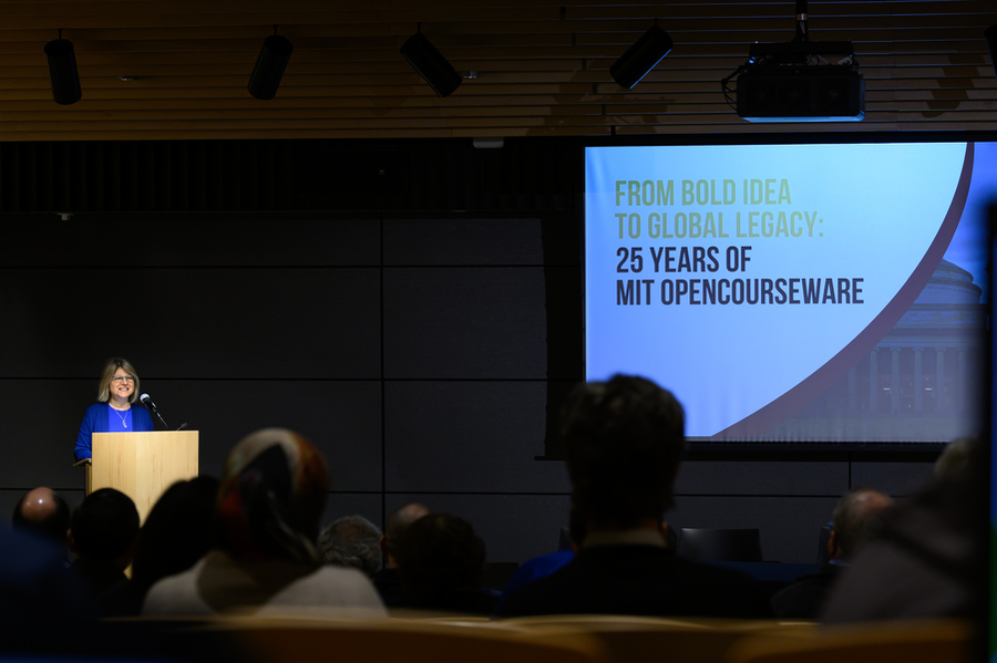 A woman wearing a blue top speaks to an audience, with a slide in the background that says "From Bold Idea to Global Legacy: 25 years of MIT OpenCourseWare"