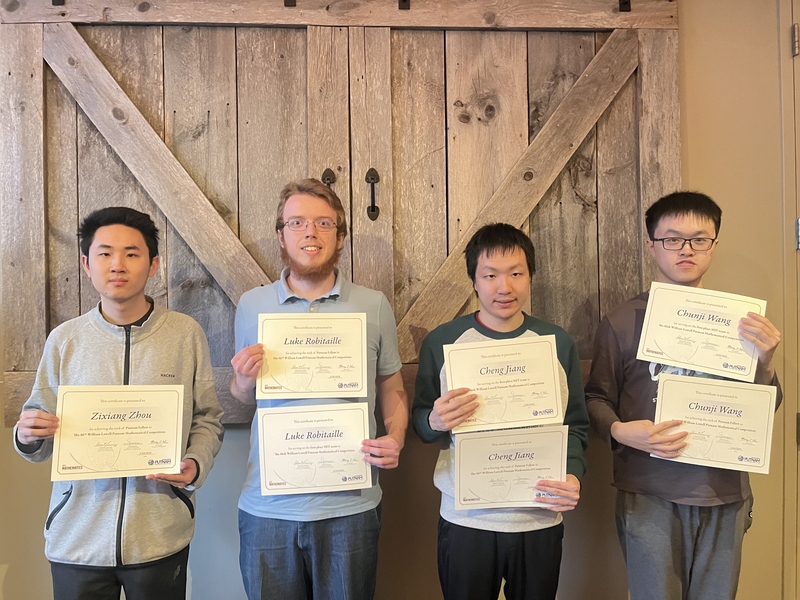 Four students pose in front of a pair of rustic wooden doors. Each is holding up either one or two certificates bearing his name.