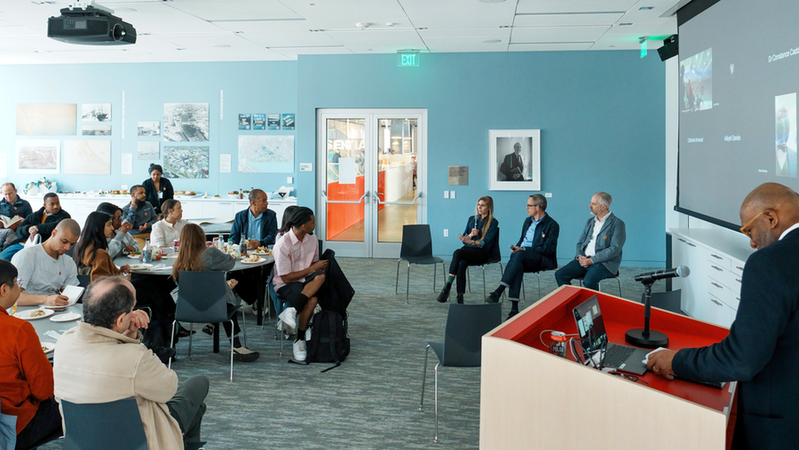 15 people sit at tables, listening to a panel of three in a conference room. A Zoom chat room with other attendees is projected at right