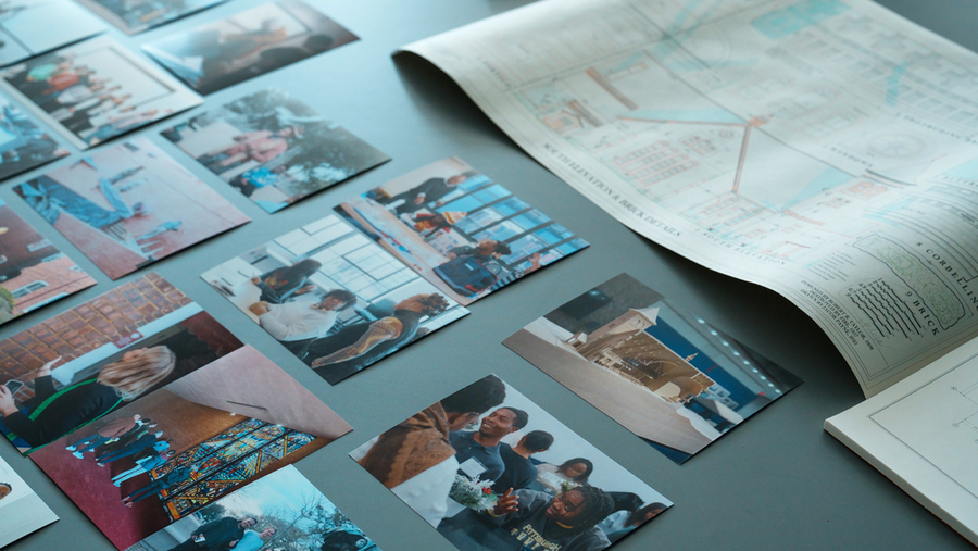 Printed color photos spread across a table show students and faculty engaged in activities and visits at MIT and Tuskegee University, alongside an architectural drawing