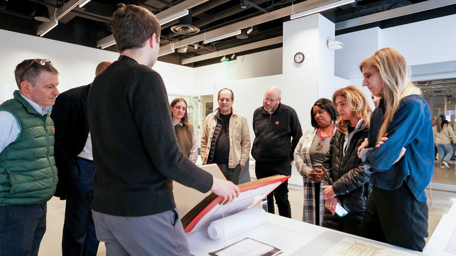 About a dozen students and faculty stand around a table at the MIT Museum as a presenter opens a large archival book