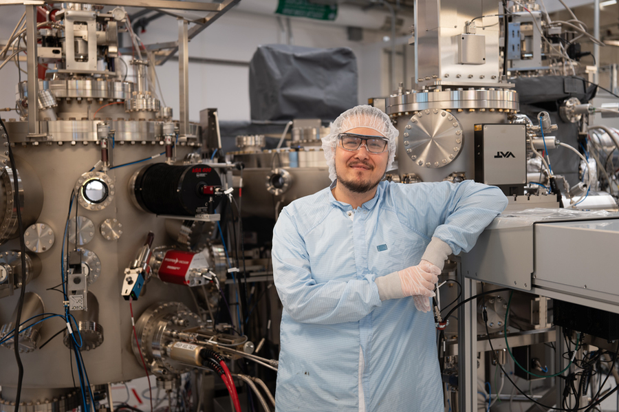 Patrick Strohbeen, in cleanroom garb, poses in with large cyllindrical tanks covered with many valves and sensors