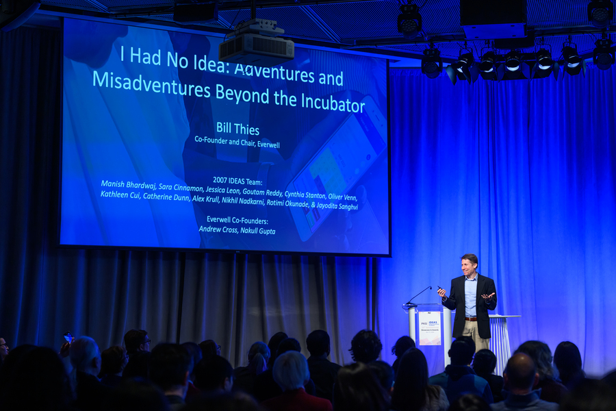 Bill Thies speaks on an indoor stage under a screen showing his keynote title: "I Had No Idea: Adventures and Misadventures Beyond the Incubator"