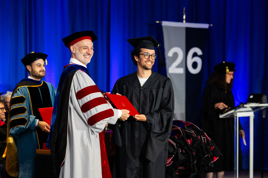 A graduate receives their diploma on stage and poses with faculty member in regalia.
