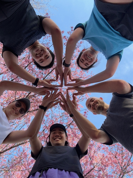5 students standing above a camera, looking down into the lens while joining their hands together to create a star. Blue sky and trees with blooming florals in the background overhead.