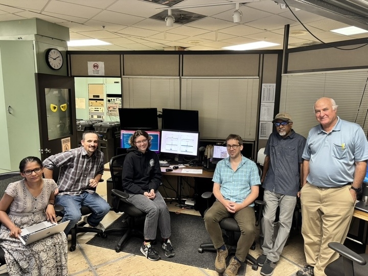 Six people pose with a computer and several monitors in a small office