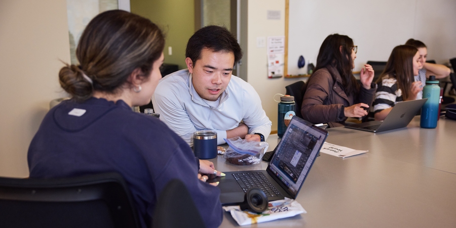 Patrick Yeung, sitting at a table, discusses information on his neighbor's laptop, with three other students in the background doing the same.