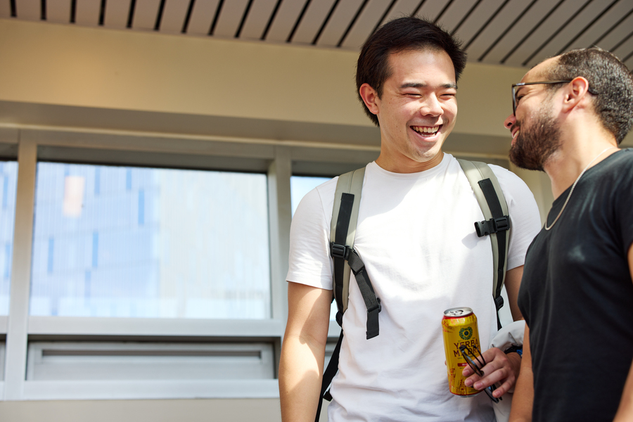 Two students laughing and chatting in a hallway, with a bright window in the background