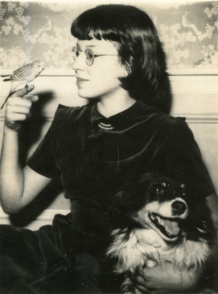 Sepia toned image of a young Harriet Latham with a parakeet perched on her right index finger, and her left arm around a happy-looking dog