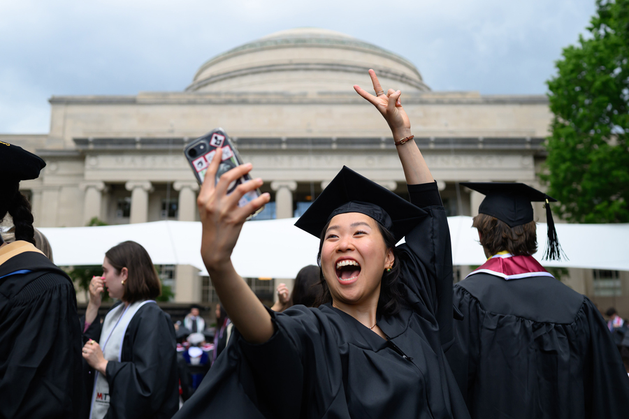 A female student in cap and gown takes a selfie in front of the Great Dome