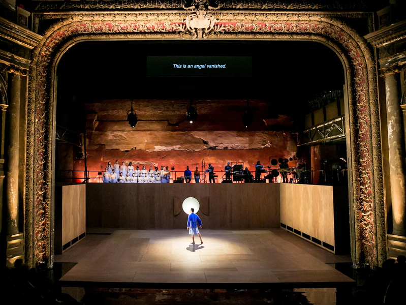 The stage design features a golden arch opening on a giant stage with bare wooden walls. A single dancer performs in a spotlight. On top of the walls are musicians against a rough rocky background.