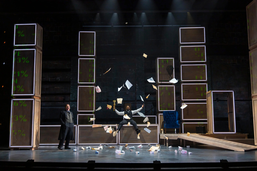 Two men in formal wear sing while one throws trash and paper in the air. The stage is mainly constructed with large rectangular boxes that have green typography projected onto them.