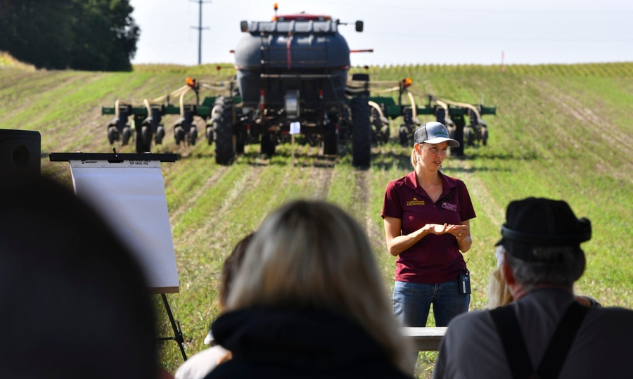 Nora Hertel stands in front of a tractor in a field, speaking to listeners in the foreground.