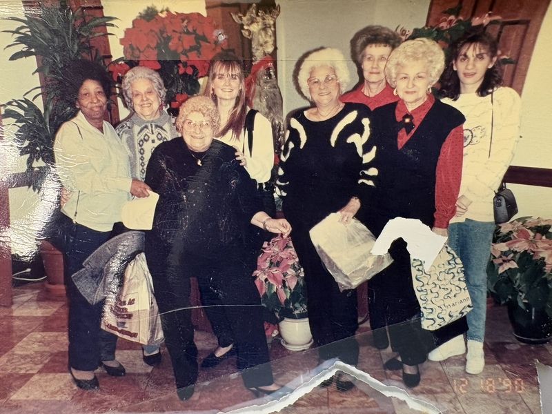 1990 photo of 8 women posing indoors. Various pointsettias are all around them.