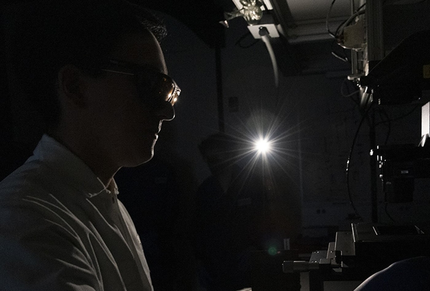 Silhouetted close-up profile of a lab member, lit from behind, glancing through a mircoscope in a darkened room.