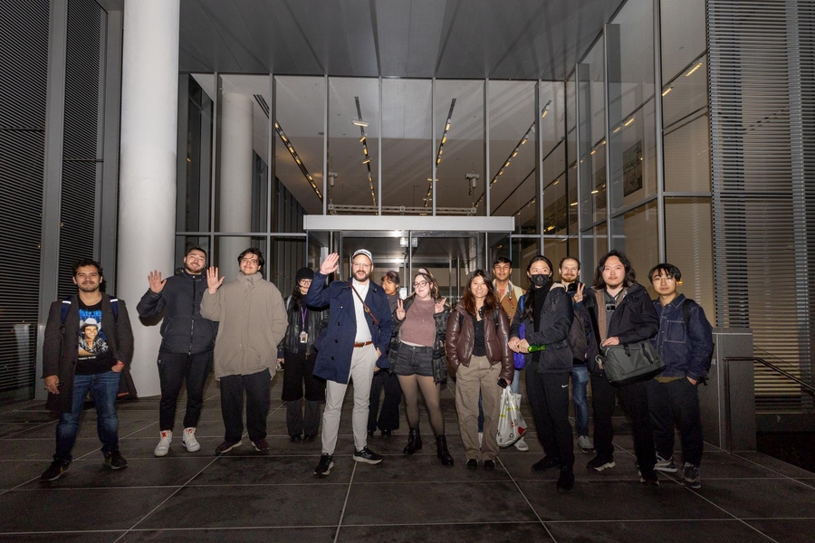 13 people pose near an entryway. Three are waving at the camera and two are flashing the two-finger peace sign.