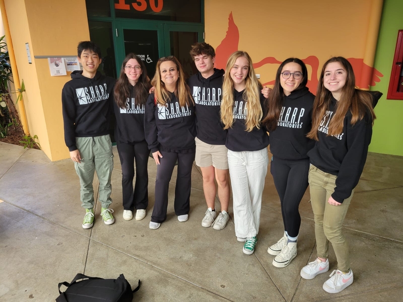 6 high school students flank their teacher outside of a yellow building. All wear sweatshirts that say "Sharp Laboratories."