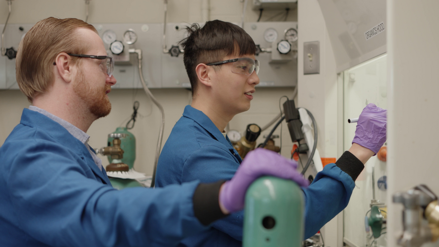 Glen Junor and Fang-Yu Kuo, wearing blue lab coats and safety glasses, work in a wet lab