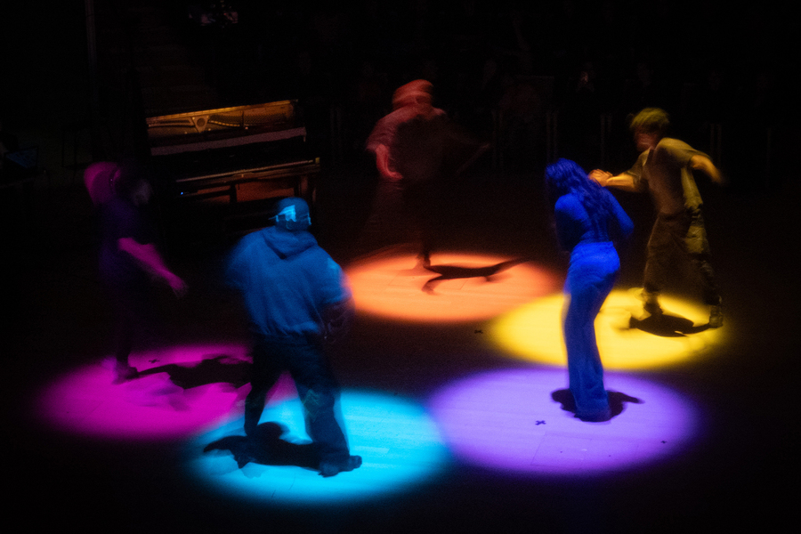 Five people blurred by motion stand in a circle on a stage, each illuminated by different colored spotlights—yellow, blue, purple, and pink—with a piano visible in the background.