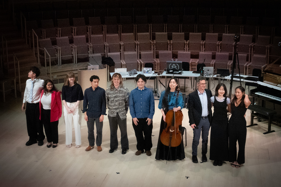 10 individuals stand in a line in front of a piano. Behind them are tables bearing electronic equipment, and a bank of empty seats