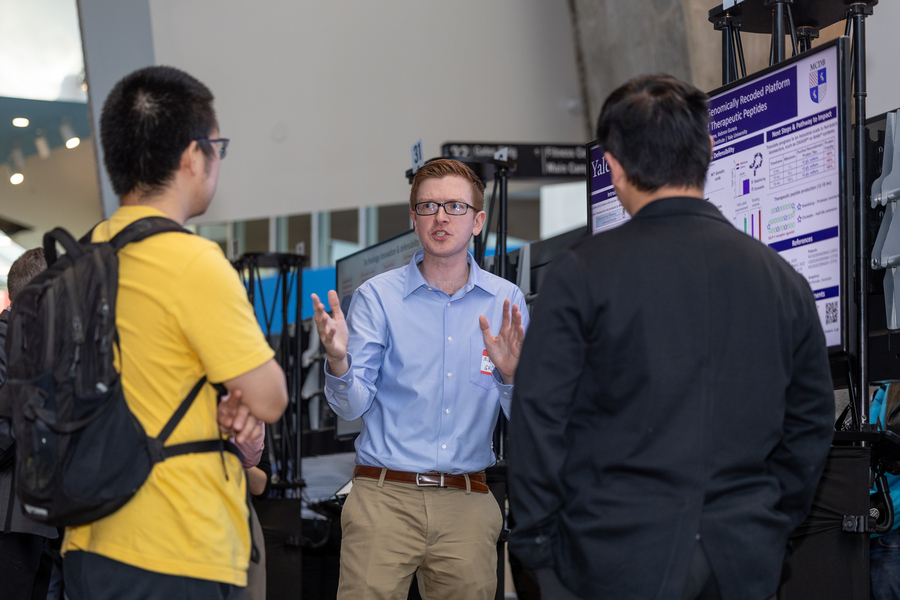 A researcher explains his poster with two attendees whose backs are to the camera