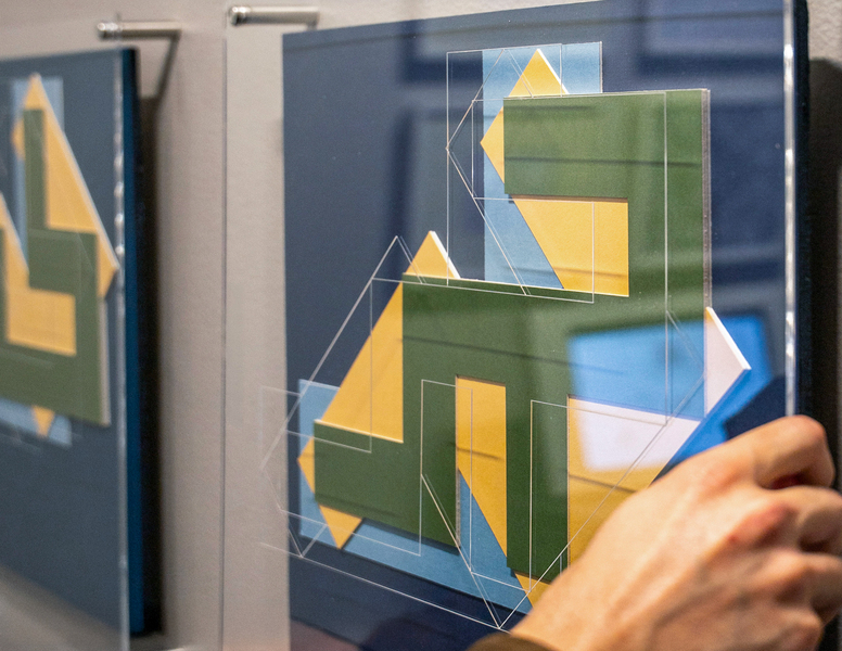 A hand manipulates a sheet of plexiglass over an interactive installation, showing layered transparent acrylic panels with geometric forms that visualize computational relationships