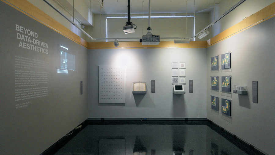 Wide view of a modest-sized room featuring wall-mounted prints, archival materials, digital displays, and geometric compositions