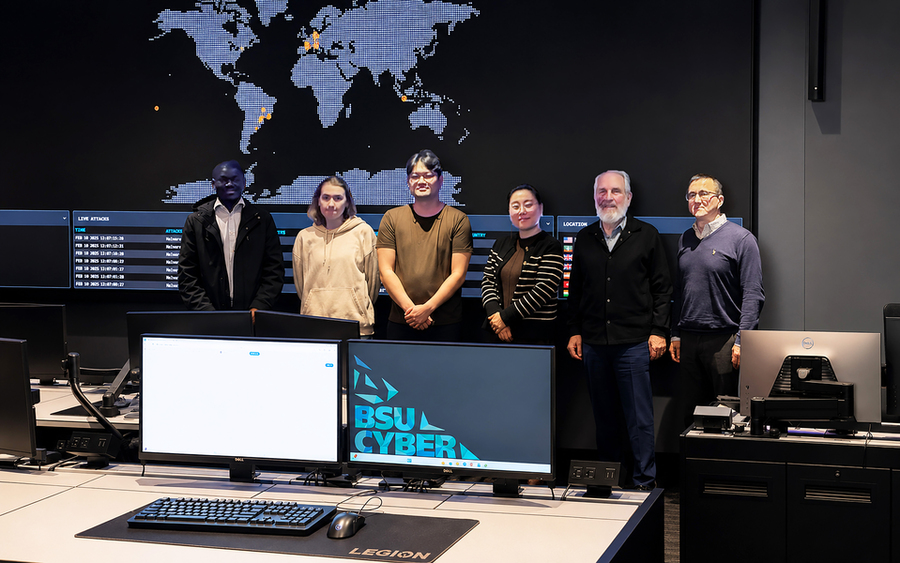 Six people pose standing in front of a wall-sized digital world map. Two computer screens dominate the foreground. One is blank and the other displays “BSU CYBER"