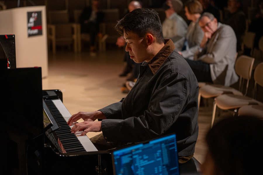 Andrew Li plays a piano on an indoor stage, with several others behind him