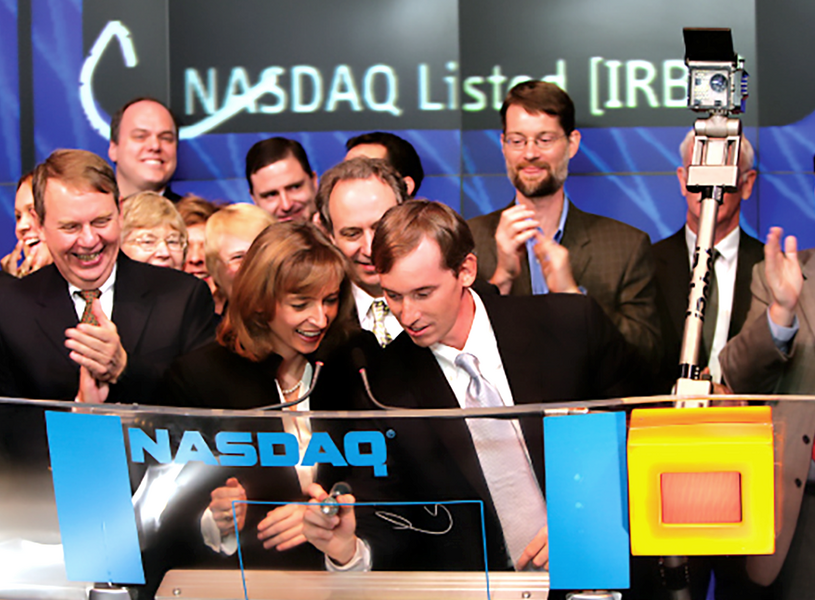 Numerous people in business attire cluster around a clear glass featuring the NASDAQ logo