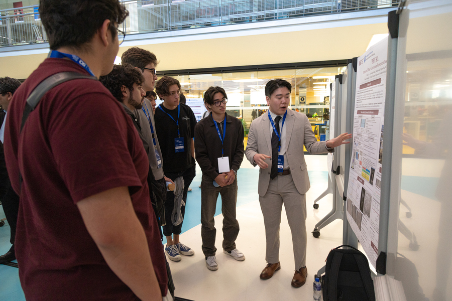 Six standing students look at a research poster while its creator explains it