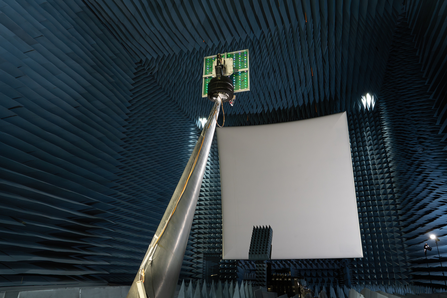 A square antenna, seen from behind, is held up toward a reflector, high above the floor, in an anechoic chamber. 