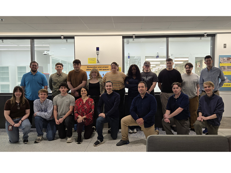 18 people pose in two rows in front of MIT.nano's cleanroom.