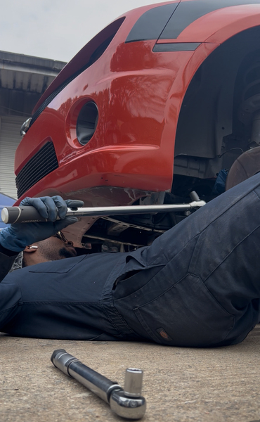 Morgan lies on his back underneath the front bumper of a bright red Camaro. He is holding a socket wrench. 