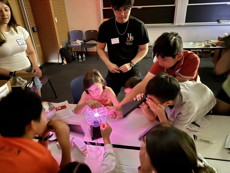 Half-a-dozen children cluster around a plasma sphere as two MIT students watch