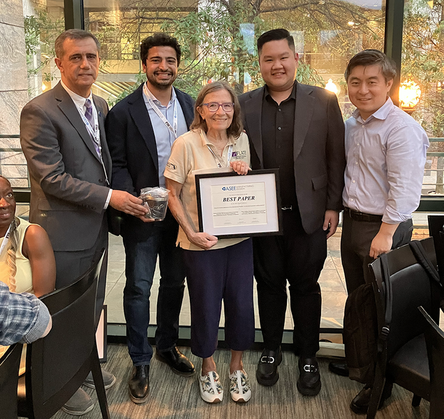 Five people pose in front of a large glass window. The person in front is holding a paper award.
