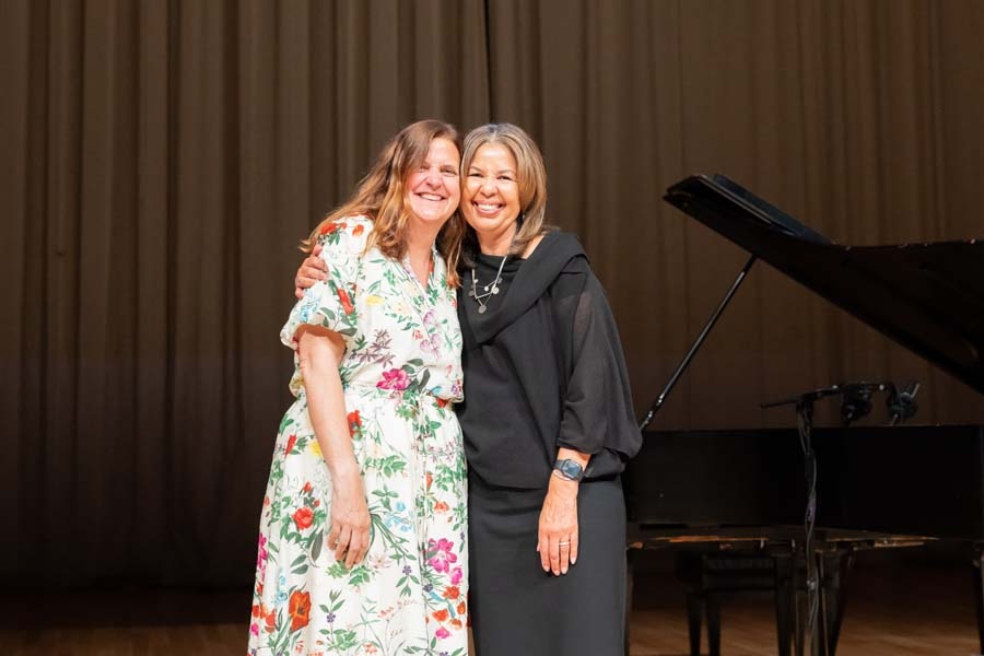 Kate McCarthy and Ramona Allen, smiling with their arms around each other on stage in front of a grand piano. 