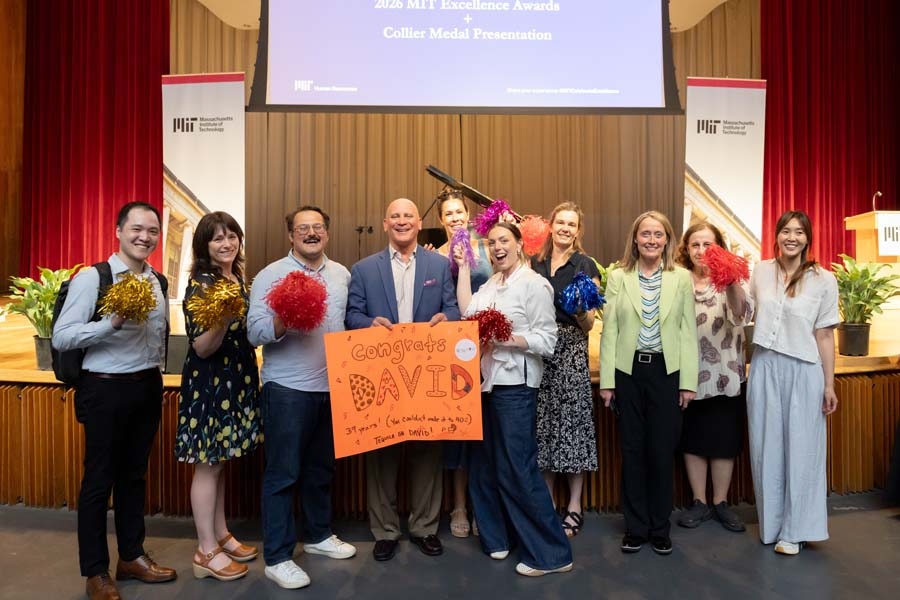 10 people pose on stage. David Verrill holds an orange paper congratulatory sign, while his colleagues and friends each hold a pompom.