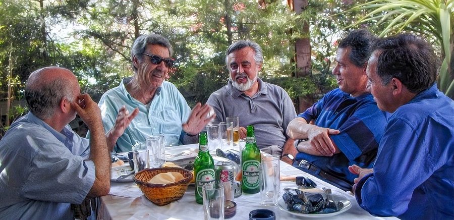 Bertsekas sits flanked by four other men of similar age at an outdoor table shaded by trees. On the table, glasses of beer and a basket of bread are strewn.