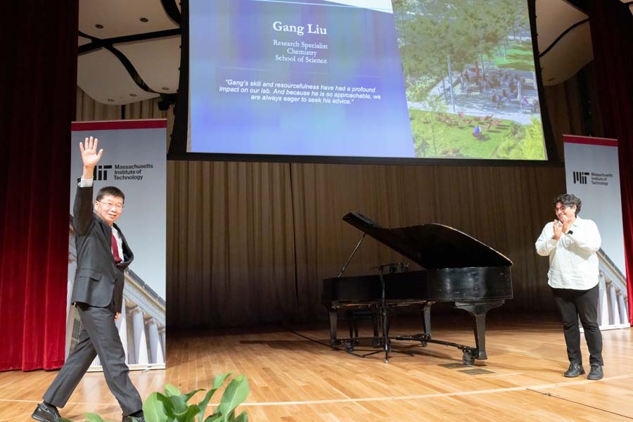 Gang Liu waves to the audience while Nergis Mavalvala claps on the auditorium stage. A screen displays information about honoree Gang Liu.