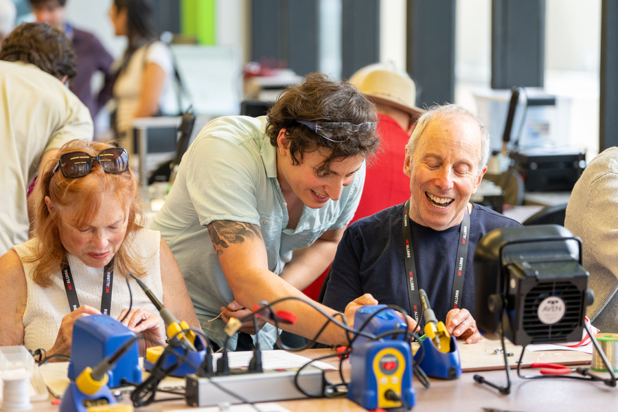 Anthony Pennes leans over the shoulders of two alumni, gesturing at the components on an electronics bench.