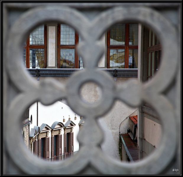 In the foreground, an ornately carved piece of gray stone holds four round openings in a cloverleaf pattern. Through the openings, a Florentine streetscape is visible, with buildings and windows reflecting the sky's light.