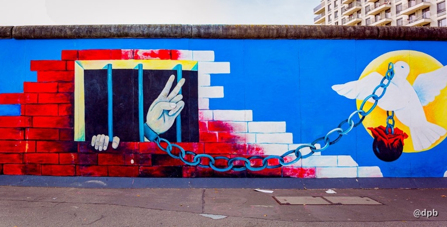 A colorful mural on a tall brick wall shows a shackled hand giving the two-finger “peace” sign from behind a barred window. To one side, a white dove carries the end of the chain connecting to the hand.