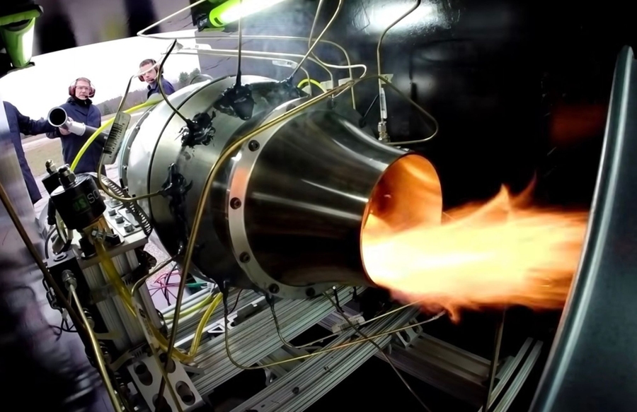 A jet engine producing waves of flame is seen from inside a test chamber. Two researchers stand at the front of the chamber.