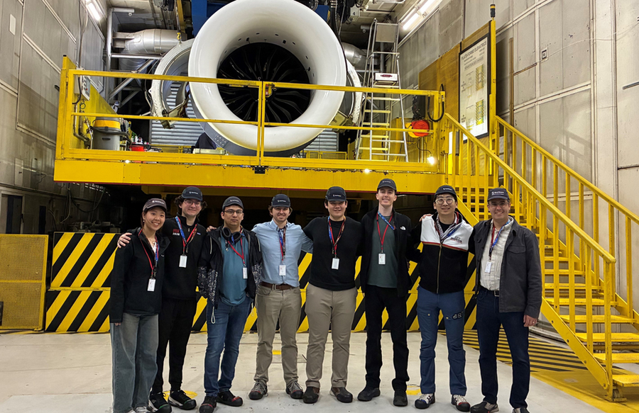 A line of 8 people stand on a machine shop floor wearing hard hats. A jet engine is behind them on a metal platform.