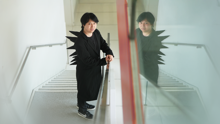Pat Pataranutaporn stands on a staircase wearing a black coat with dramatic fabric spikes on his back, his reflection visible in a glass panel beside him.